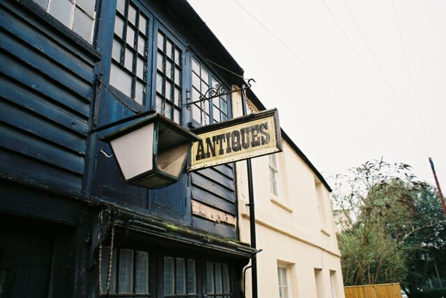 A film photograph of an antiques shop. A sign reading 'Antiques' hangs perpendicular to the building aside a lantern which has a broken fascia.
