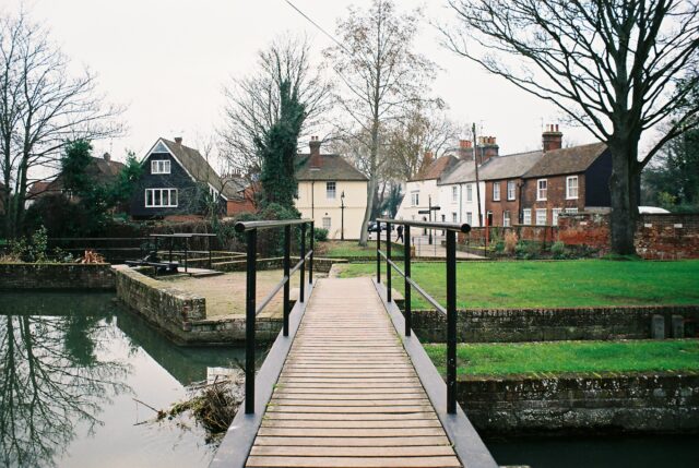 Looking from one side of a short bridge to the other. The bridge spans a small channel of water and features wooden slats and black iron handrails.