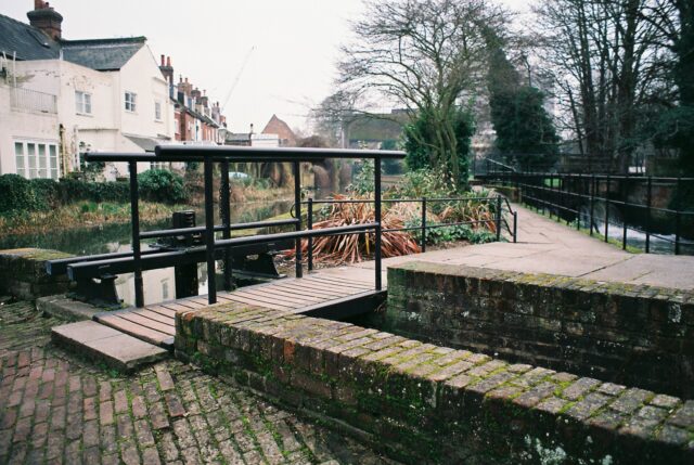 A short bridge over a small channel of water with wooden slats and black iron handrails.