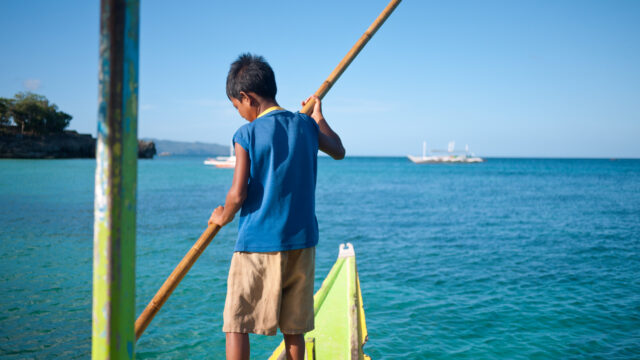 Taken from inside a boat, this image shows a young boy stood on the vessel's bow holding a stick to guide the boat.