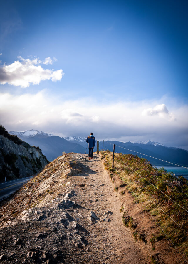 Image of a man walking up a small dirt mount with their back to the camera. In the background, beautiful mountains and a lake a visible.