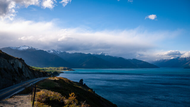 Landscape image of a large lake with mountains in the background. On the left a road runs around the lake.