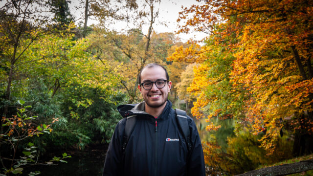 Photo of myself by the waterside at Winkworth arboretum with yellow and green autumnal trees behind me. The lakehouse is just about visible in the background.