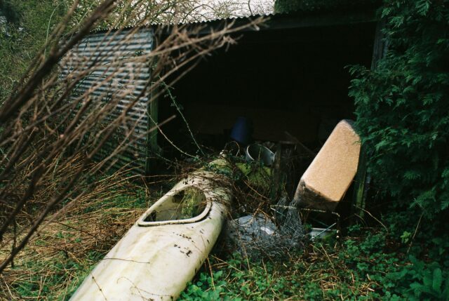 A shed structure with corrugated metal panelling set amongst undergrowth. The shed door is open and various discarded items are visible. A kayak, which appears unused for many years, protrudes from the shed opening.