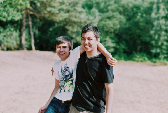 Two brothers stood in a sandy area with woodland behind them. The younger brother has his arm wrapped around the older one and appears to almost be 'hanging' off his sibling.