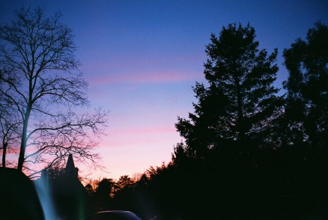 A sunset photograph featuring a tree and an oast house in silhouette against a purple, red and blue sky.