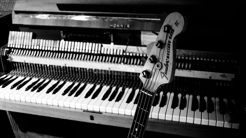 Black and white photo of a Hohner piano missing its front panel such that the internal components are visible. The headstock of a Fender Mustang bass guitar is in the foreground.