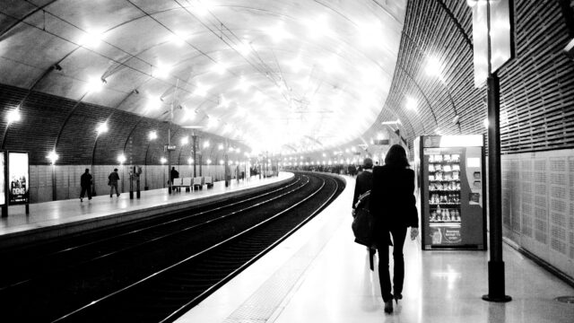 Black and white image looking down the curved platform of Monaco Monte Carlo station. In the foreground, a female commuter is walking towards a mass of commuters, further down the platform. The scene is brightly lit with several white lights on the station roof.