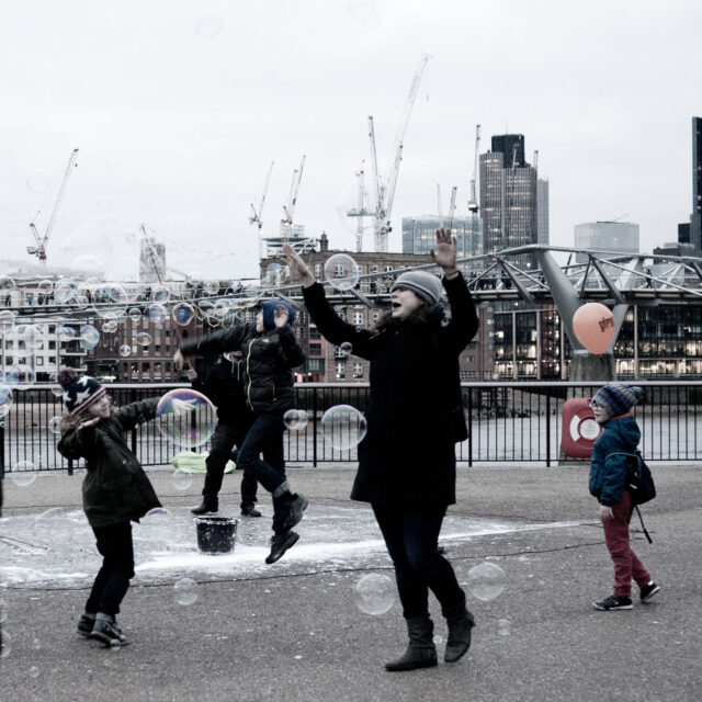 On the South Bank in London a street entertainer has blown lots of bubbles. two children and one adult are jumping in the air popping the bubbles, whilst a third child, holding an orange balloon, is stood looking at the fun.