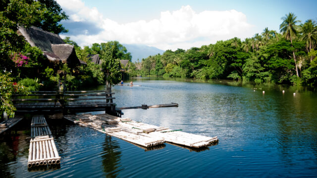 Looking up a river at a plantation. In the foreground a jetty and bamboo rafts are floating, and in the background are huts with wooden roofs and trees lining both sides of the river bank.