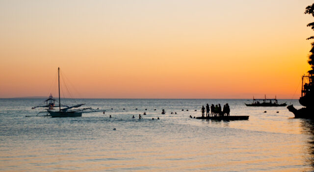 A view out to sea at sunset. In the foreground, silhouetted, there are boats, buoys and a pontoon with people on it. There are more people swimming in the water having dived off the pontoon.