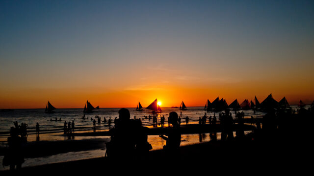 Looking out from a busy beach on a golden and blue sunset. In the background, silhouettes of several sailboats are visible and in the foreground, silhouetted people are all over the beach enjoying the view.