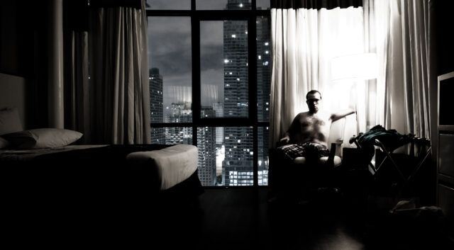 Image of a man sitting in a hotel room chair illuminated by a lamp. Also visible is the bed, and the view from the window of a cityscape. The photo is almost colourless and moody.