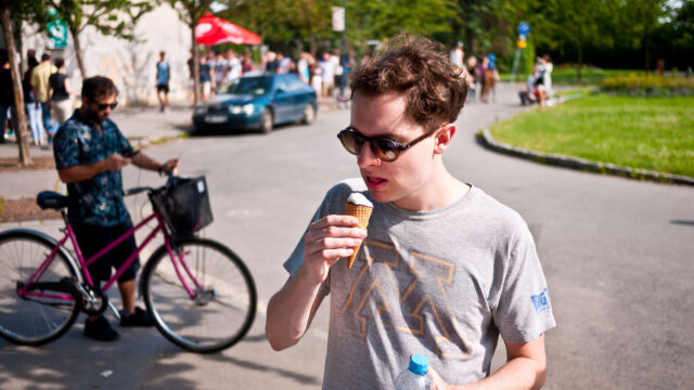 A man eating an ice cream. In the background is a garden and a different man in shadow with a bike