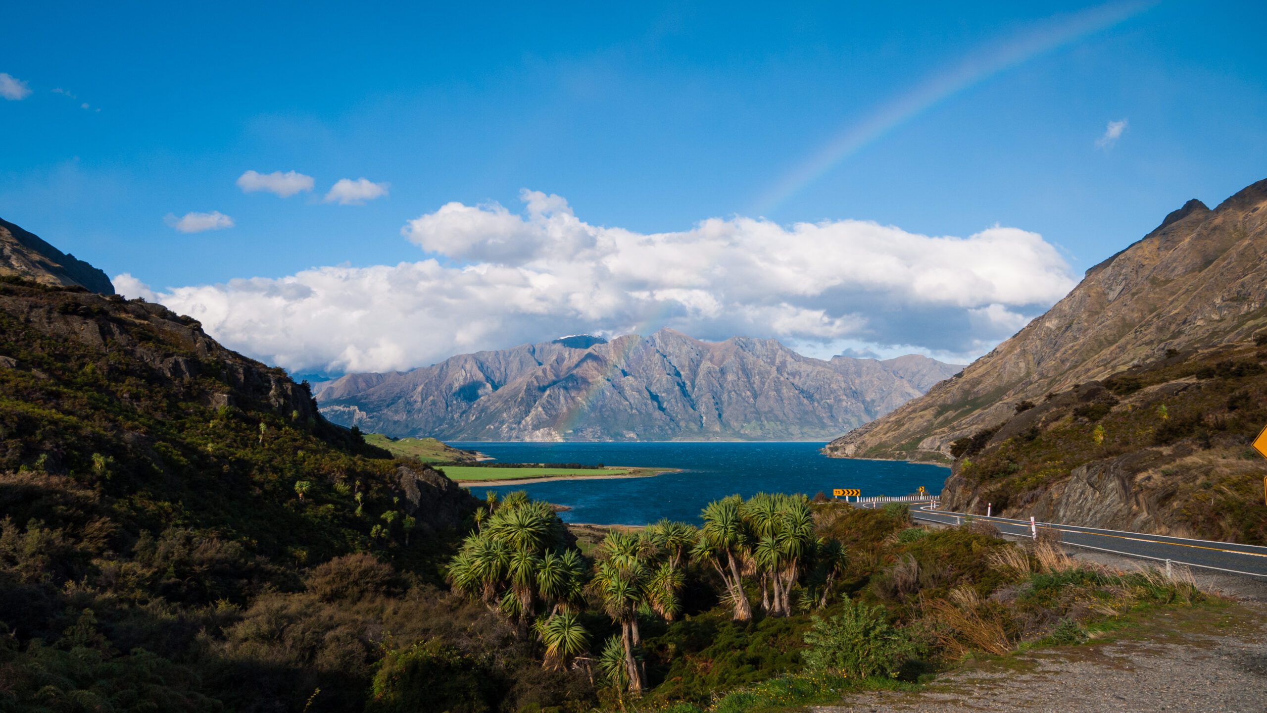 Image showing a view over a large lake with a mountain in the background in New Zealand. The scene is sunny and there is a rainbow arcing from the right of the picture over the blue lake.