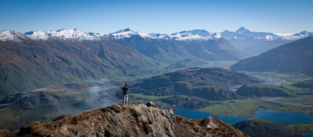 Landscape image taken from the top of a mountain. A mountain range and lake are visible in the background and the image is vibrant blue. In the center of the image there is a mountain ridge jutting out and a man is visible walking on the ridge and surveying the view.