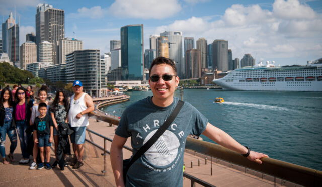 A man standing in Sydney Harbour with the business district skyscrapers in the background. To the man's right is a family of six who have ended up in the photo inadvertently whilst posing for their own shot.