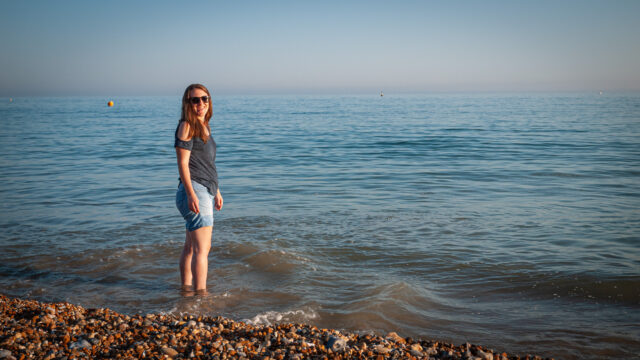 A woman standing in the shallows of the sea on a pebble beach at dusk, looking back towards the camera.