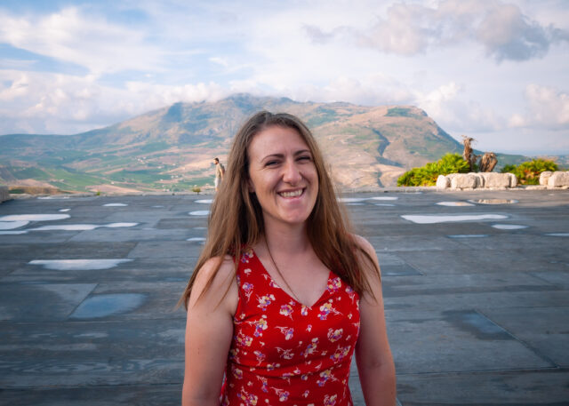 A portrait shot of Annika wearing a red summer dress whilst stood on the stage at the Segesta theatre with a mountain in the background. It is a sunny evening.