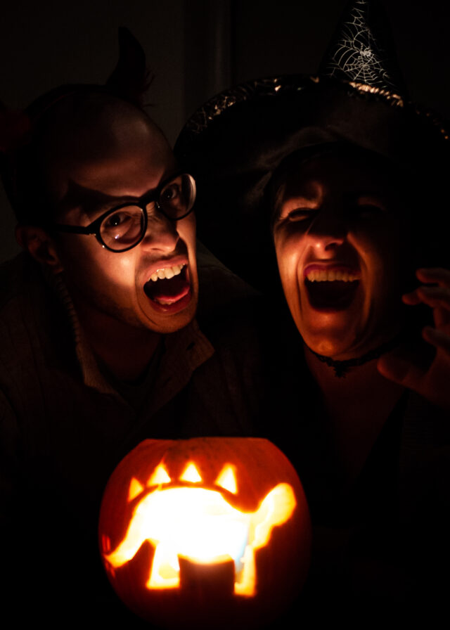 Darkly lit image of a man and a woman illuminated from below by candlelight from a pumpkin carved with the image of a stegosaurus.
